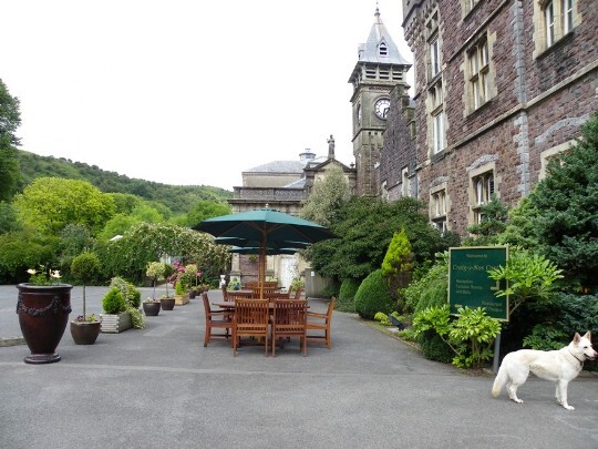 Garden seats and tables with green canopies line the front of the castle, bordered by flower and tree tubs and barrels Craig y Nos Castle Wedding Venue in South Wales Front Courtyard showing clock tower and garden tables and chairs